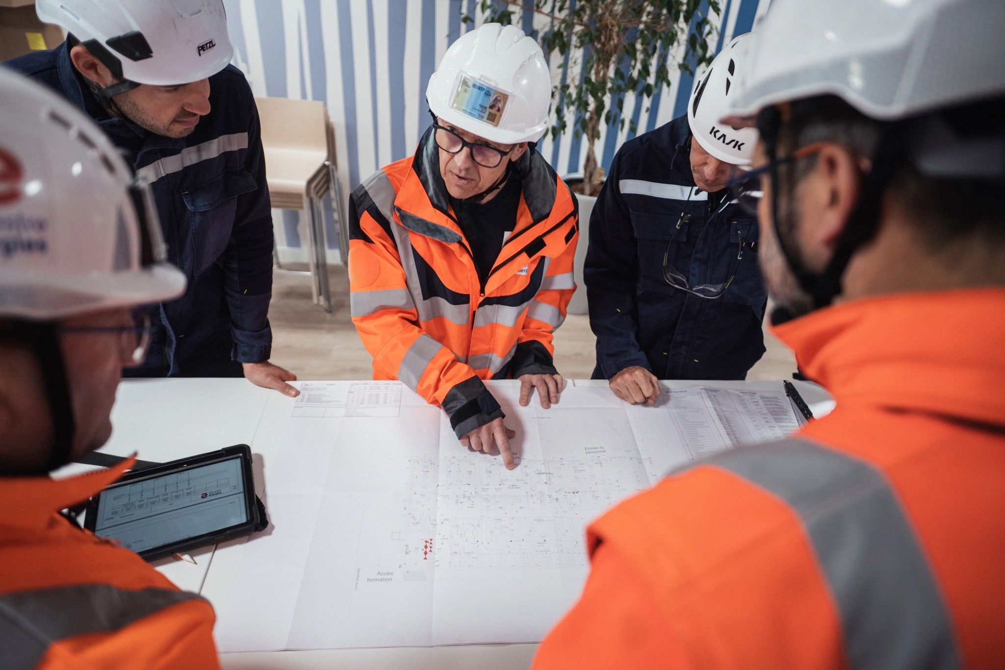 Cinq techniciens en gilets haute visibilité et casques de sécurité analysent un plan technique sur une table, illustrant la planification de projets en électricité tertiaire, sécurité et transition énergétique.