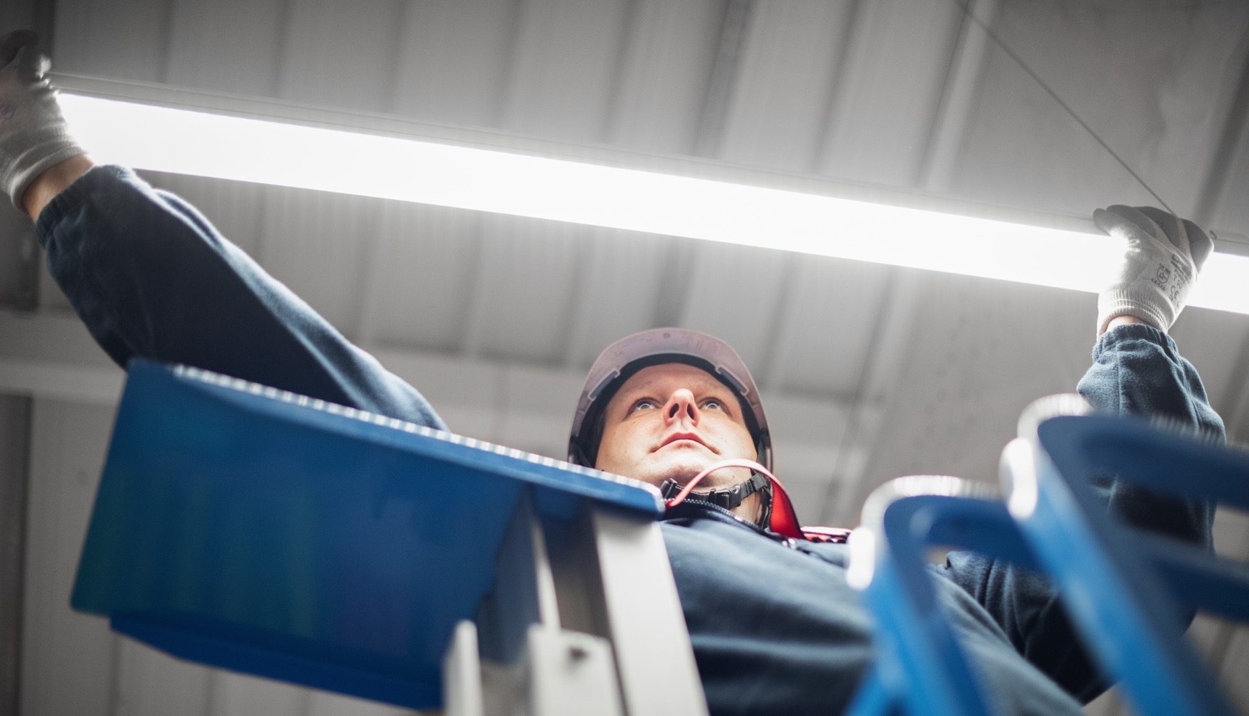Une personne en tenue de travail, vue de dessous, est en train d'installer ou de réparer un tube fluorescent au plafond.