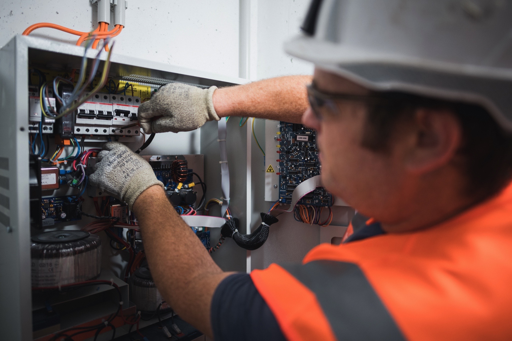 Technicien portant un gilet de sécurité orange et des gants, effectuant des travaux sur un panneau électrique contenant des fils et des composants électroniques.