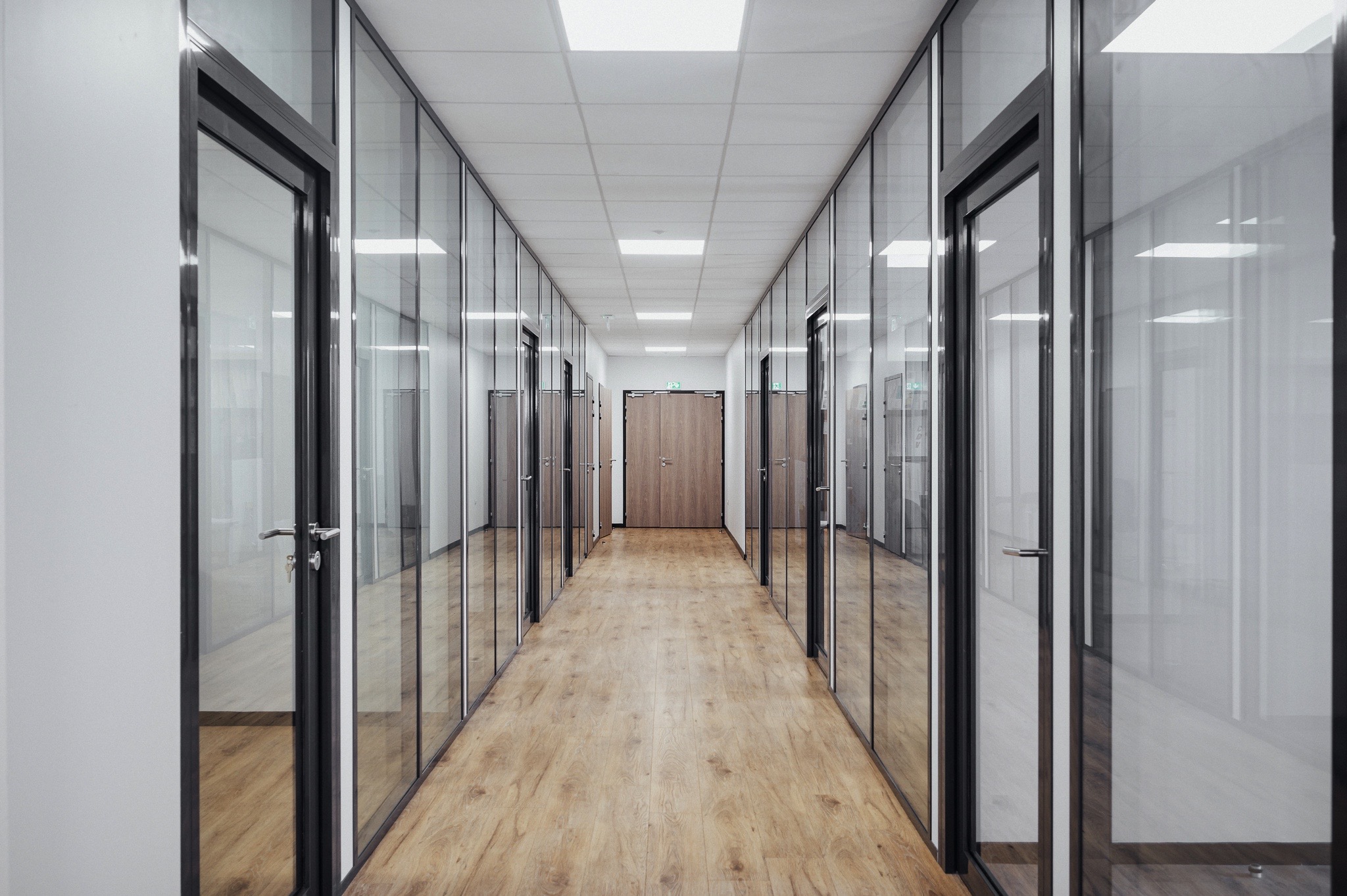 Un couloir de bureaux modernes avec des cloisons vitrées, un sol en bois clair et un éclairage encastré.