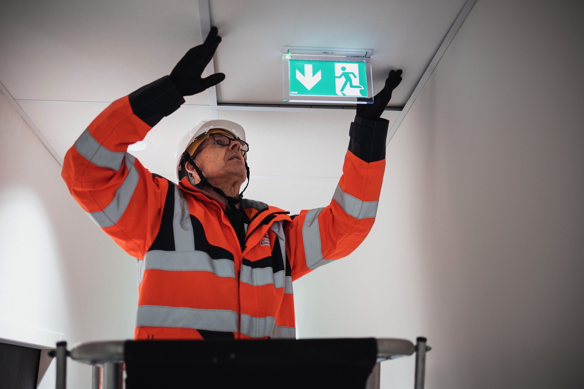 Technicien en haute visibilité installant un panneau de sortie de secours, illustrant les services d’électricité tertiaire, de sécurité et d’éclairage LED proposés par Sylvestre Énergies.