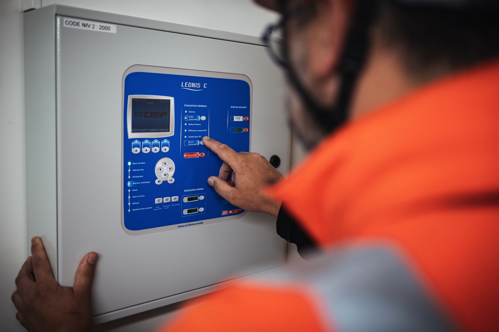 Technicien en veste orange interagissant avec un panneau de contrôle LEONIS C, utilisé pour la gestion des alarmes et de l’évacuation dans les bâtiments tertiaires, illustrant les services de sécurité et de maintenance de Sylvestre Énergies.
