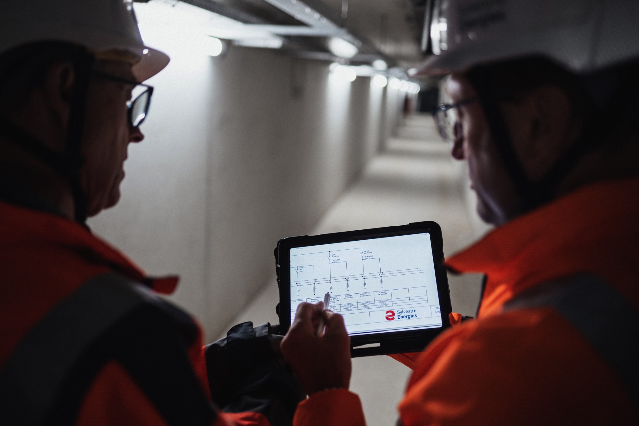 Deux techniciens en veste de sécurité orange et casque consultent un plan technique sur tablette dans un couloir, illustrant les services de maintenance, d’électricité industrielle et de gestion de projet de Sylvestre Énergies.