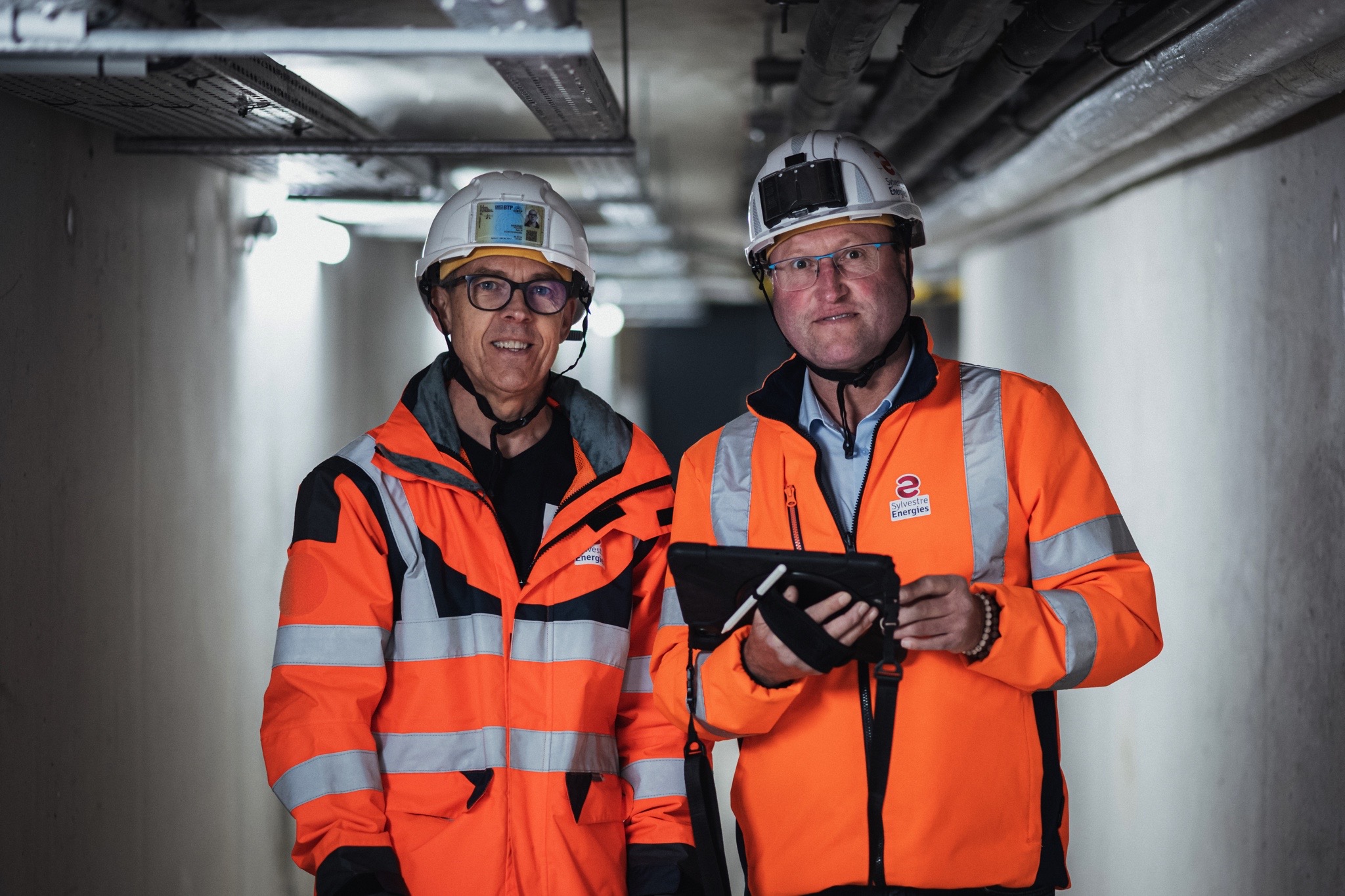 Deux techniciens en veste haute visibilité et casque de sécurité effectuent une inspection dans un couloir technique, l’un d’eux tenant une tablette, illustrant les services de maintenance, d’électricité industrielle et de sécurité de Sylvestre Énergies.