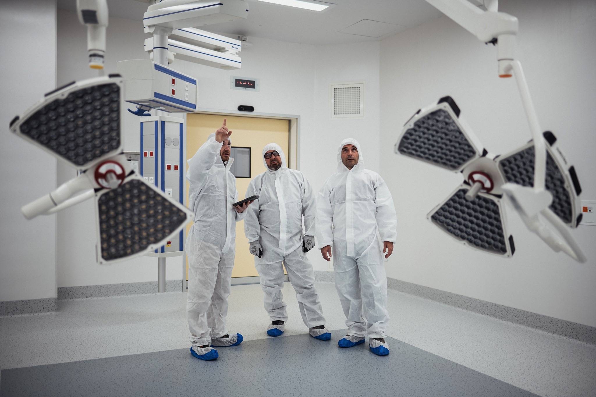 Trois personnes en tenue de protection intégrale discutent dans un environnement stérile, l’une d’elles tenant une tablette.