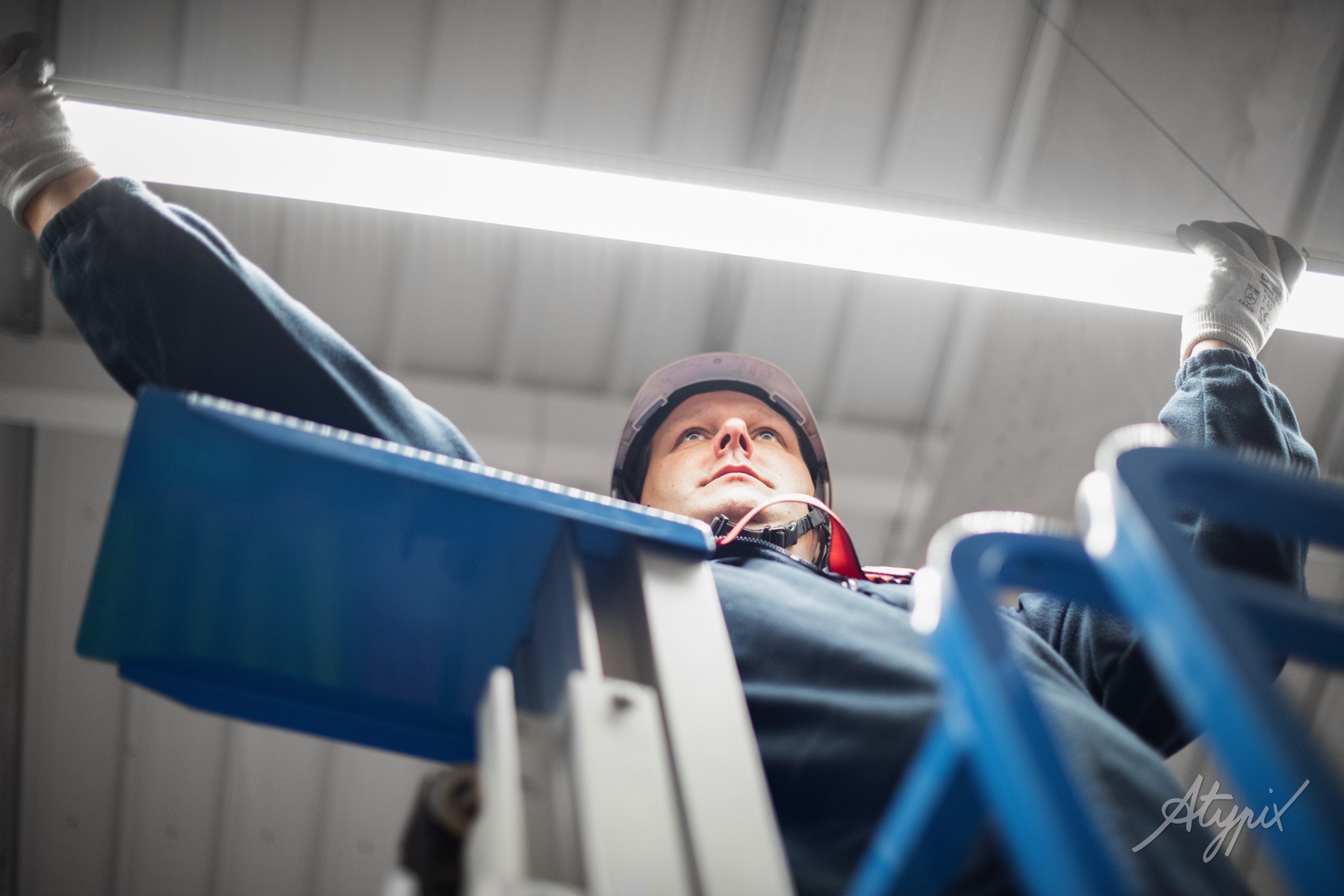 Une personne en tenue de travail, vue de dessous, est en train d'installer ou de réparer un tube fluorescent au plafond.