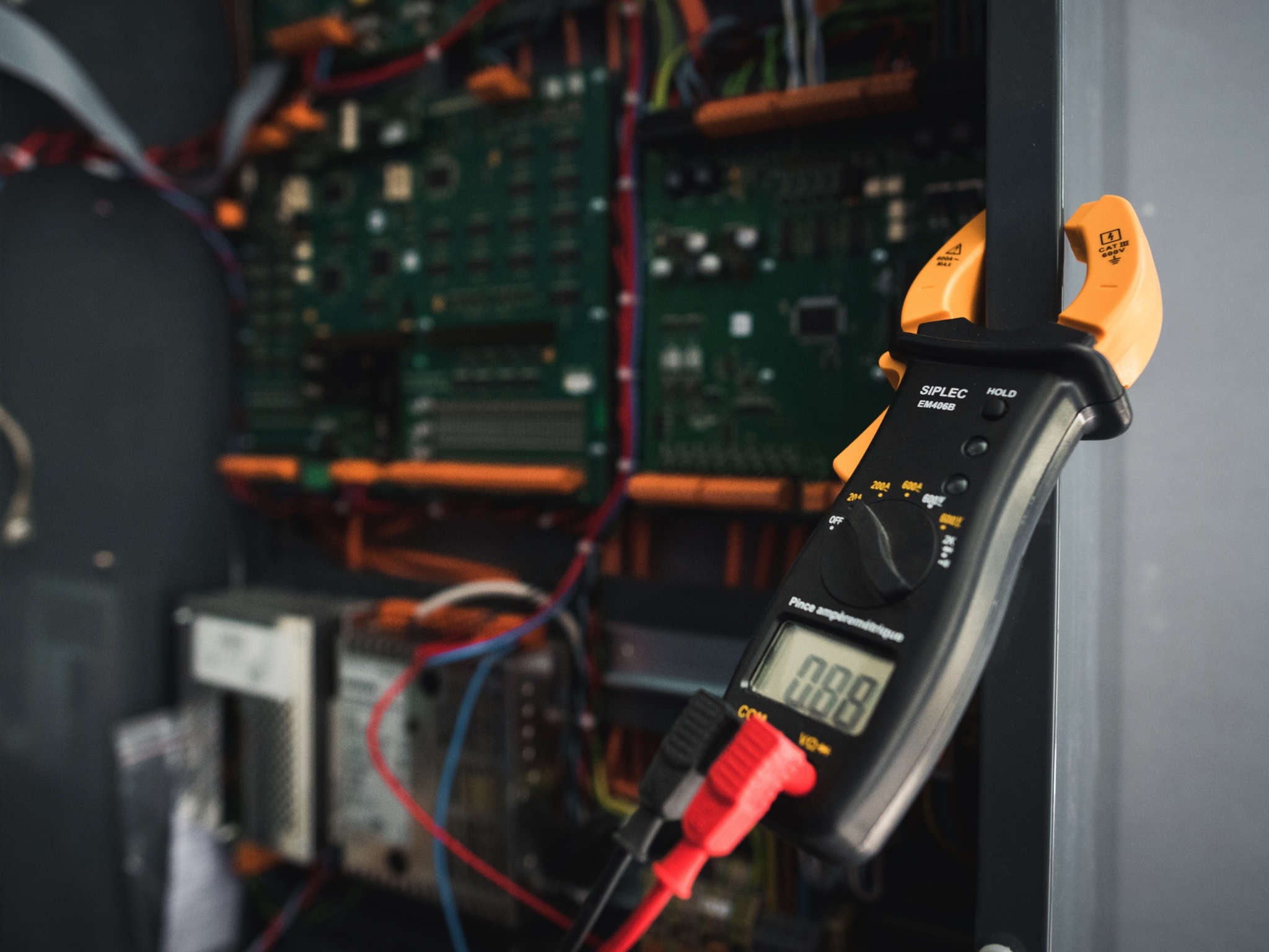 Un tableau électrique avec câblage complexe et circuit imprimé, sur lequel un technicien utilise un multimètre à pince pour mesurer la tension, illustrant les services de maintenance, d’électricité industrielle et de sécurité de Sylvestre Énergies.