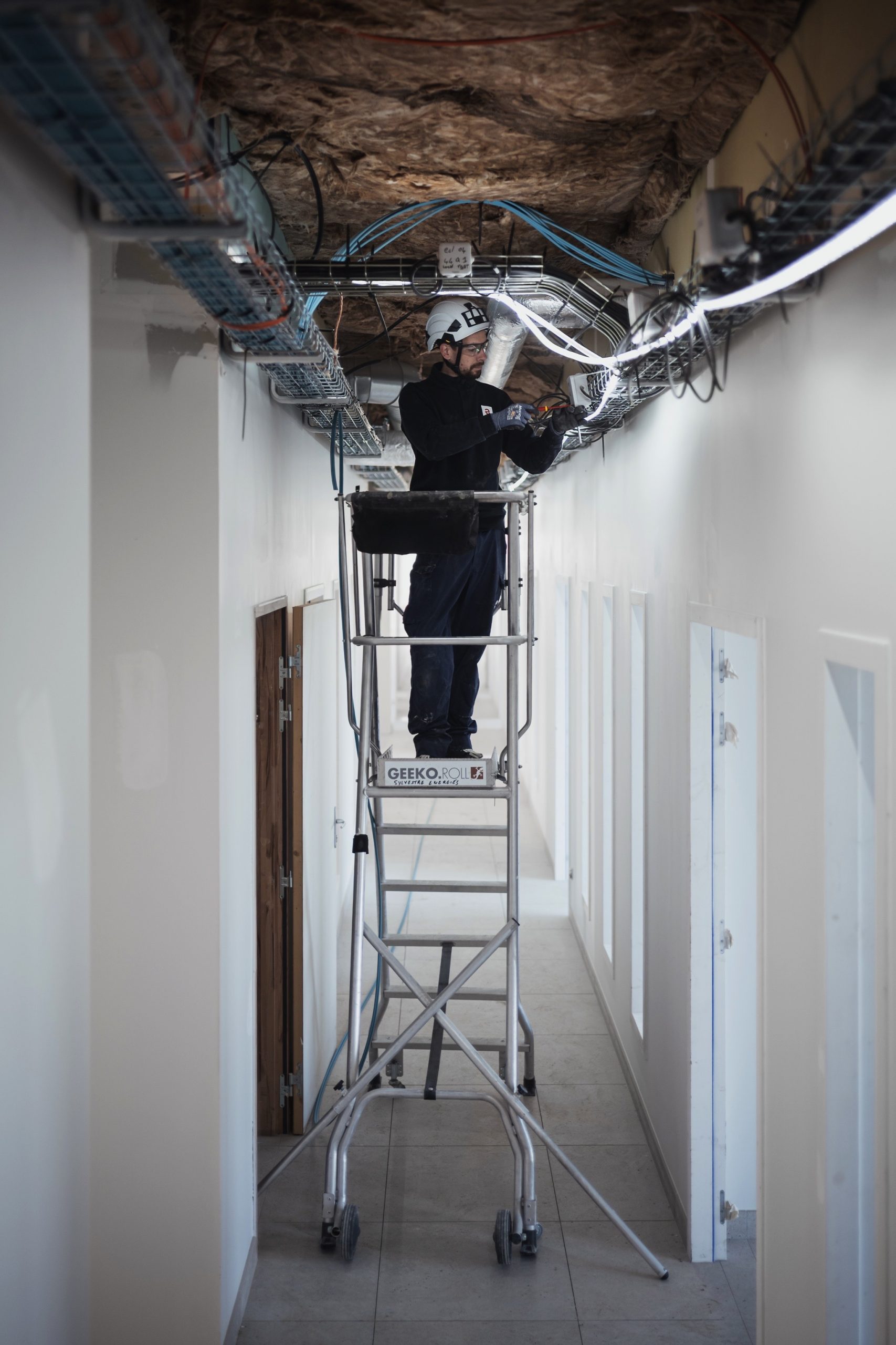 Technicien réalisant des opérations de câblage et de raccordement électrique en hauteur sur un chemin de câbles dans un bâtiment tertiaire.