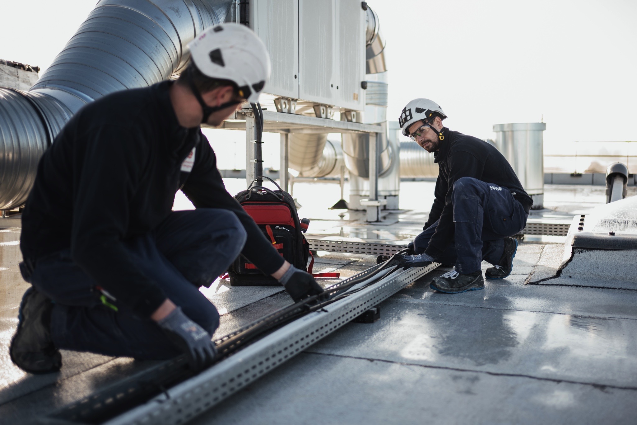 Techniciens réalisant l’installation de chemins de câbles électriques sur une toiture technique à proximité d’équipements de ventilation et de traitement d’air.
