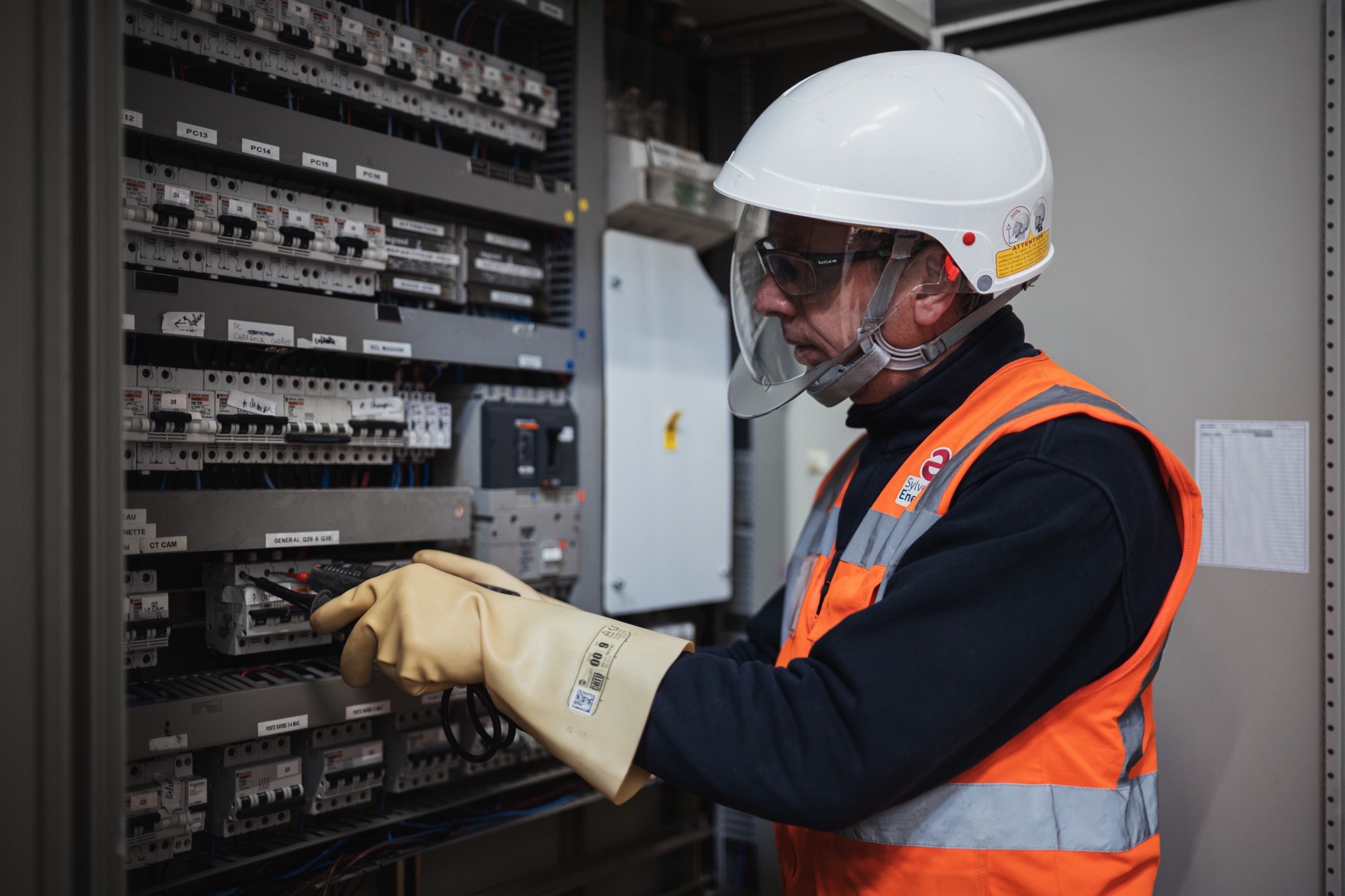 Technicien équipé d’EPI réalisant des mesures et vérifications sur une armoire électrique dans le cadre d’une opération de maintenance et de contrôle.