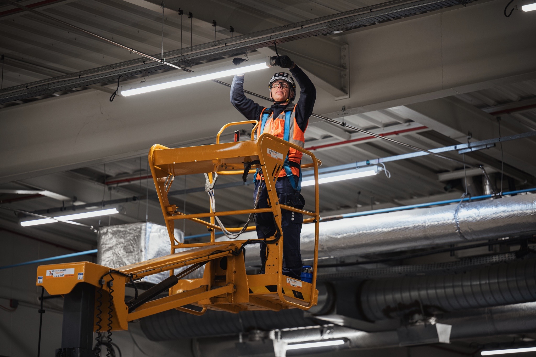 Technicien intervenant sur une nacelle élévatrice pour l’installation et la maintenance d’un système d’éclairage dans un bâtiment industriel.